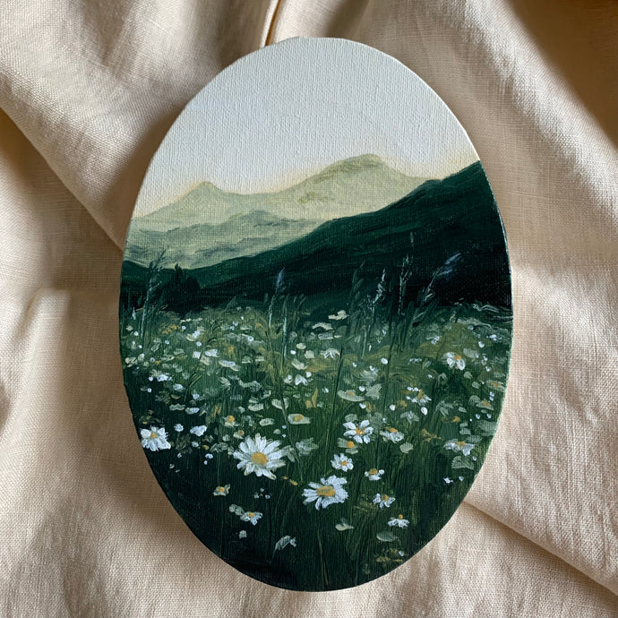 An oval painting done in oils laying on cream linen fabric. The painting has a white sky with light green mountains in the background. In front is a dark vibrant green hill which turns into a field of lighter green that is dotted with wheat and grasses and daisies like sparkles in the dark setting. It looks as though the setting sun has thrown most of the scene into shade but is catching some the little white flowers. 