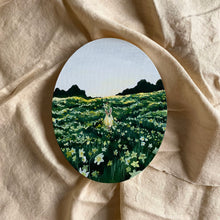 Load image into Gallery viewer, There is an oil painting done on an oval canvas laying on cream linen fabric. The painting is of a field of daffodils and in the middle of the field is a girl wearing light yellow holding a bouquet of flowers. The sky is light blue and the background trees are very dark green with the field a lighter green scattered with a variety of narcissus
