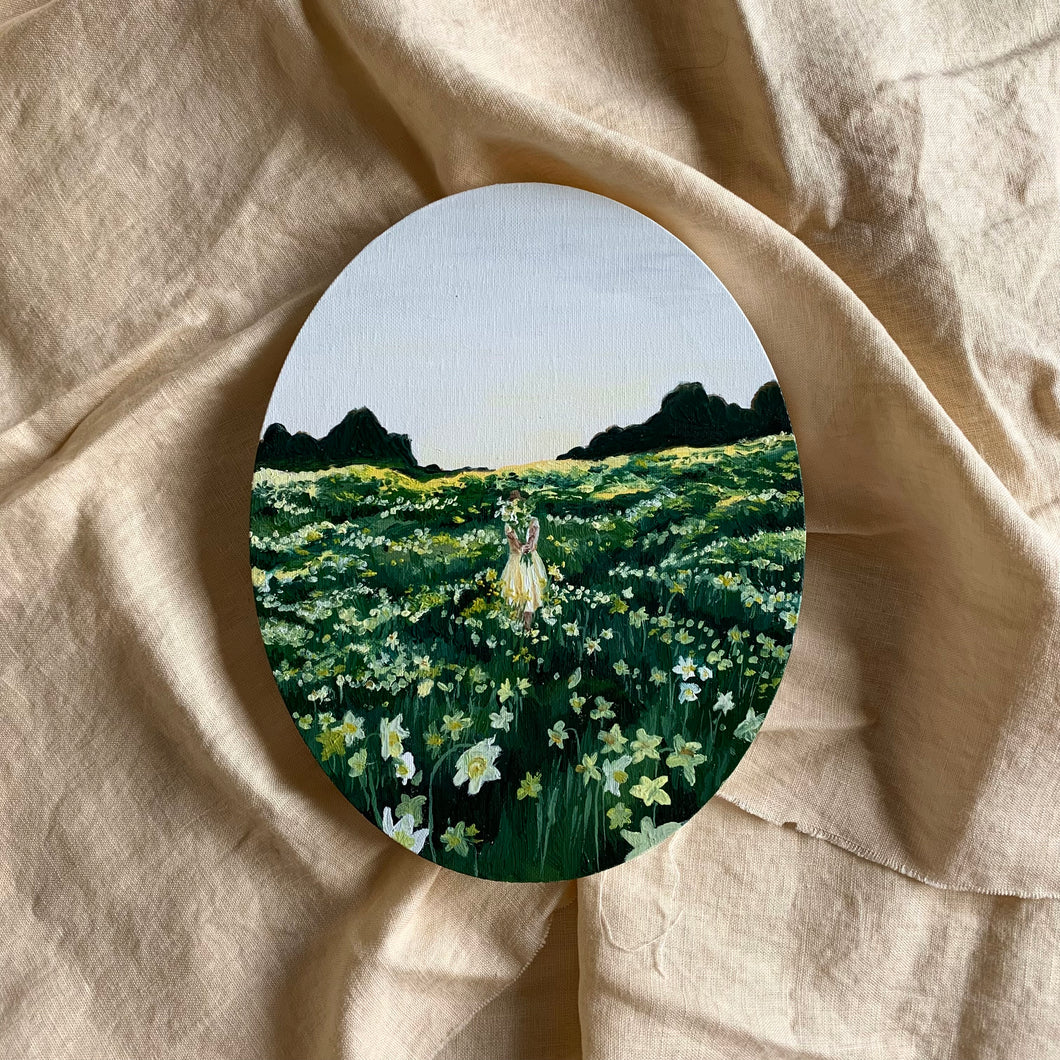 There is an oil painting done on an oval canvas laying on cream linen fabric. The painting is of a field of daffodils and in the middle of the field is a girl wearing light yellow holding a bouquet of flowers. The sky is light blue and the background trees are very dark green with the field a lighter green scattered with a variety of narcissus