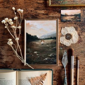 A worn table with some dried flowers and paintbrushes and an old book. All these things are showcasing the painting of the girl twirling in the field.
