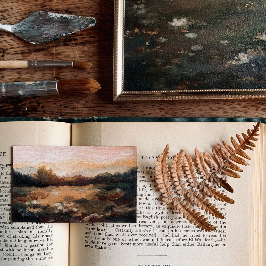 A tiny painting is resting on a book which is placed upon a worn wooden table. There is a brown dried fern on the page next to the painting that is done in oil. Some paintbrushes and a paint knife as well as a small corner of another painting in a gold frame are at the top of the photo.  The art is of a glowing sunset over some moody hills and trees. The overall feel of the piece is romantic and glowing.