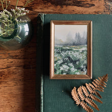 Load image into Gallery viewer, A dark green book on a worn table with a tiny painting of distant trees and a hill with a close up of white flowers. The overall painting is white, grey and green with hints of gold. 
